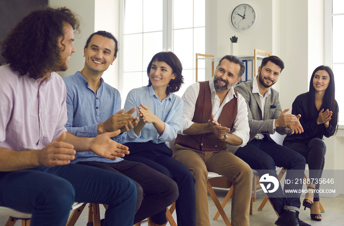 Group of happy thankful young and mature people thanking coach for interesting talk in business conference. Smiling male and female teammates sitting in row applauding creative man in team meeting