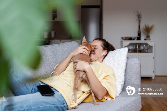 Brunette woman relaxing at home with her little dog lying on sofa hugging dog and listening music using headphones and mobile phone.