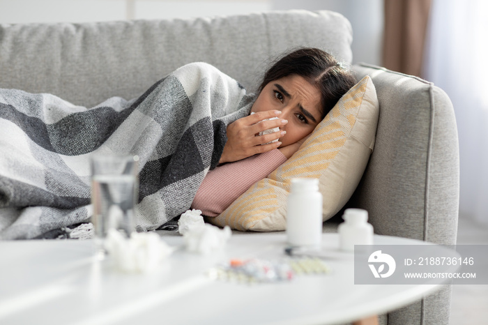 Unhappy young indian female sneezes in napkin, blows nose, suffers from fever and flu, lies on sofa