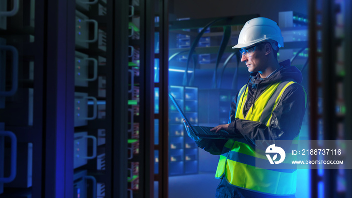 Man with laptop in server room. System administrator for industrial companies. Guy with computer. Server racks emit neon light. Maintenance industrial server equipment. Man in helmet in data center