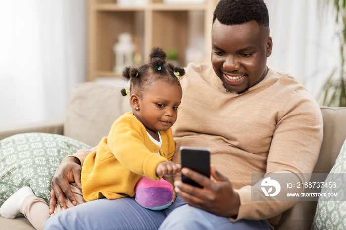 family, fatherhood and technology concept - african american father with smartphone and little baby daughter playing with ball at home