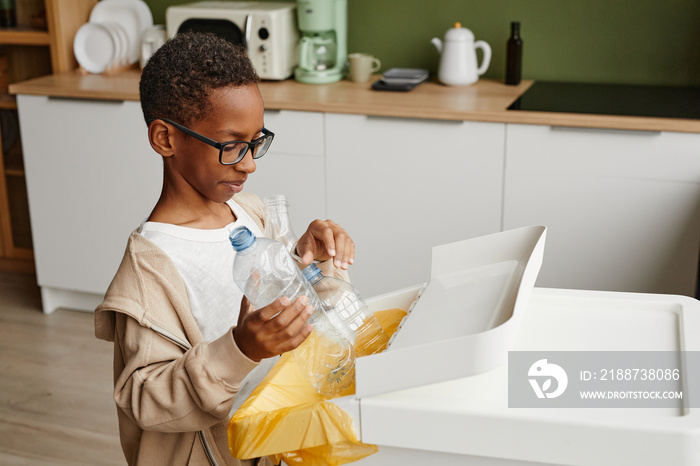 Side view portrait of African-American boy putting plastic bottles in recycling bins while sorting household waste at home, copy space