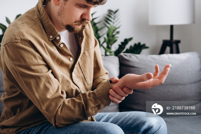 Close up of sad young man 30s suffering pain on hands and fingers, arthritis inflammation. Upset bearded male has pain in wrist sitting on couch in living room at home. Health care and medical concept