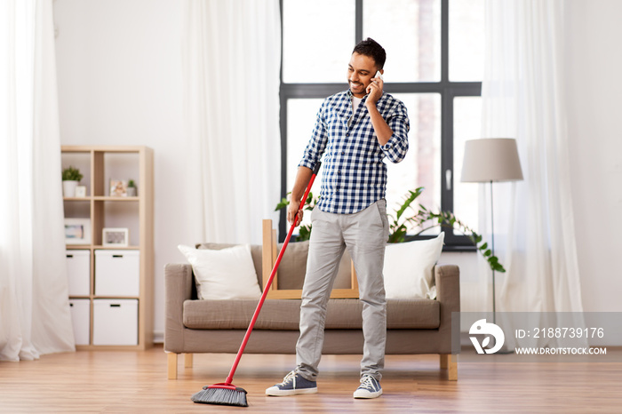 cleaning, housework and housekeeping concept - indian man with broom sweeping floor and calling on smartphone at home