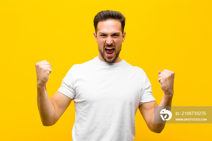 young handsome man shouting aggressively with an angry expression or with fists clenched celebrating success against orange wall