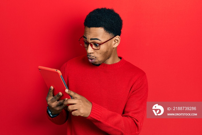 Young african american man using touchpad device scared and amazed with open mouth for surprise, disbelief face