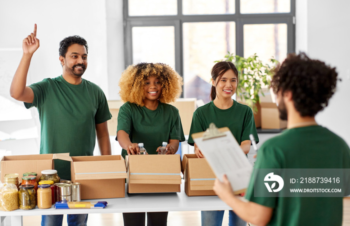 charity, donation and volunteering concept - international group of happy smiling volunteers packing food in boxes according to list on clipboard at distribution or refugee assistance center