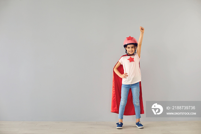 Little baby superhero in a helmet smiles raised his hands while standing against a gray background.