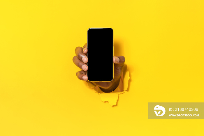 Mobile app. Closeup of black male hand holding cellphone with blank screen, breaking through yellow paper sheet