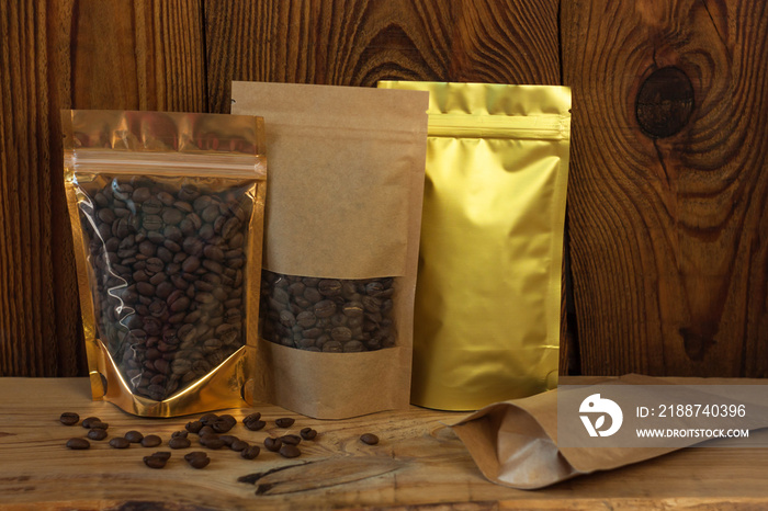 Brown kraft paper and golden metallized pouch bags with coffee beans front view on a wooden background.