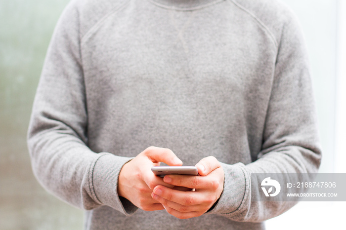 A midsection of a young male in neutral and grey colours holding a cell or mobile phone in his hands whilst texting a message.