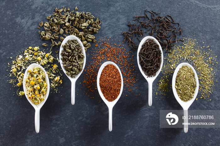 A variety of loose dried tea leaves on spoons (chamomile, green, rooibos, black and mate tea), photographed overhead on slate