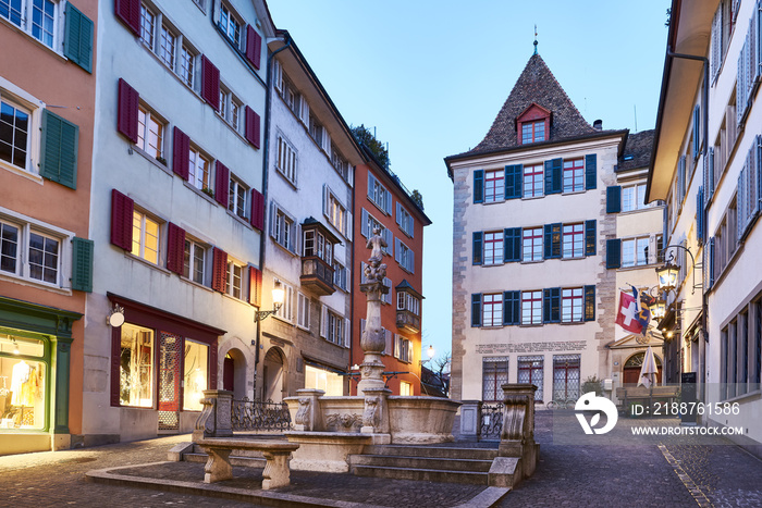 Zürich Napfbrunnen beim Napflatz an der Spiegelgasse, Abenddämmerung, Zürcher Altstadt, Brunnenturm, mittelalterlicher Wohnturm, Schaufenster, Beleuchtungen, blauer Abendhimmel, Schweizer Fahne