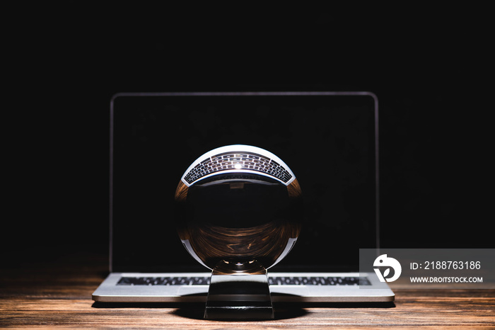 Crystal ball near laptop on wooden table on black background