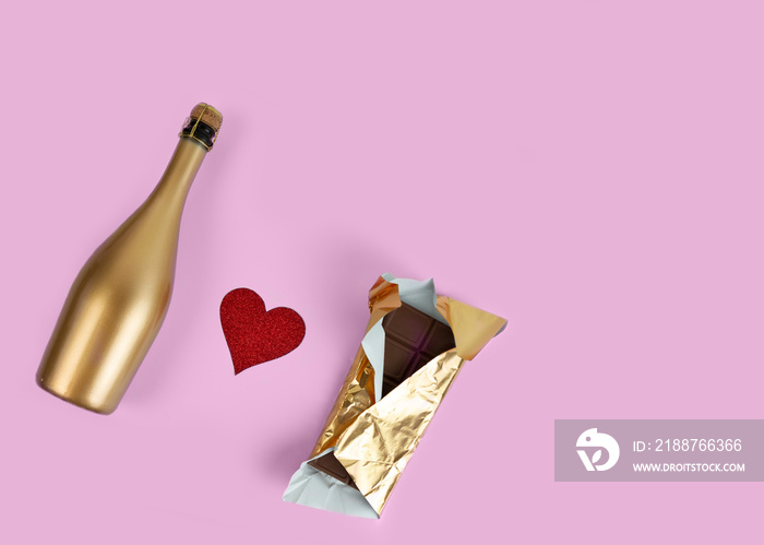 Golden champagne bottle and chocolate bar in gold wrapper and red glitter heart on a pink background. Festive set.