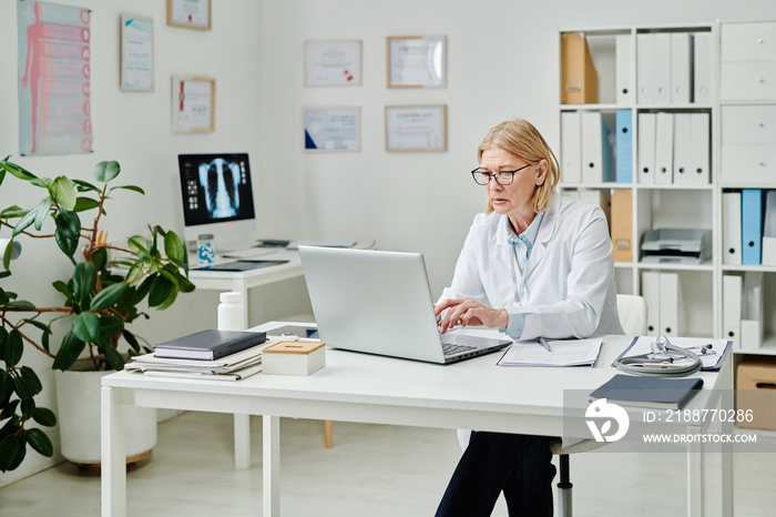 Serious mature female clinician in whitecoat answering questions of online patients while typing on laptop keyboard by desk