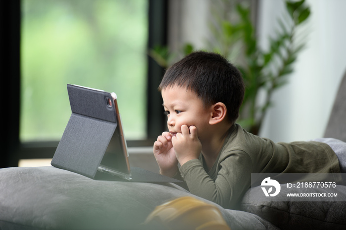 Asian boy looking at the tablet screen attentively. overstimulated children concept. Too much screen time. 3 years boy watching videos while tv is working.Smartphone, Internet addiction concept.