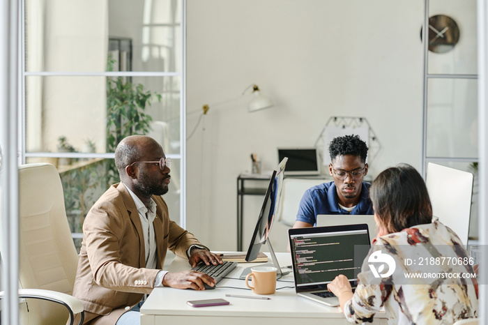 Group of programmers sitting at table and working together with new software on computers during meeting at office