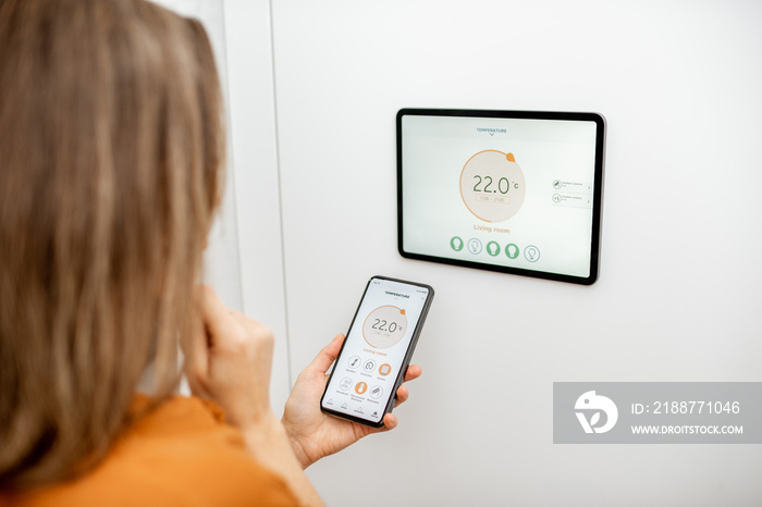 Young woman controlling temperature in the living room with smart phone and digital touch screen panel. Concept of heating control in a smart home