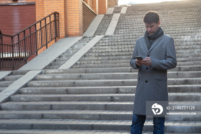 elegant man in long overcoat reads information using digital technology and standing on high steps. Concept fashion city technology