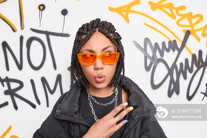 Photo of surprised female rapper with braids makes cool gesture looks wondered aside wears trendy orange shades enjoys spending time outdoor in public place poses against creative drawn graffiti wall