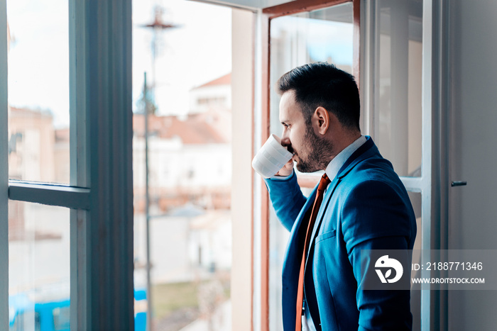 Successful business man looking at city through open window while drinking from a cup.