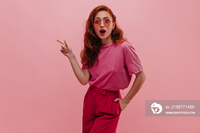 Attractive fair-skinned girl with surprise points her index finger to side on pink background. Stylish redhead woman holds hand in pocket dressed in casual pink clothes. Concept indicating copy space.