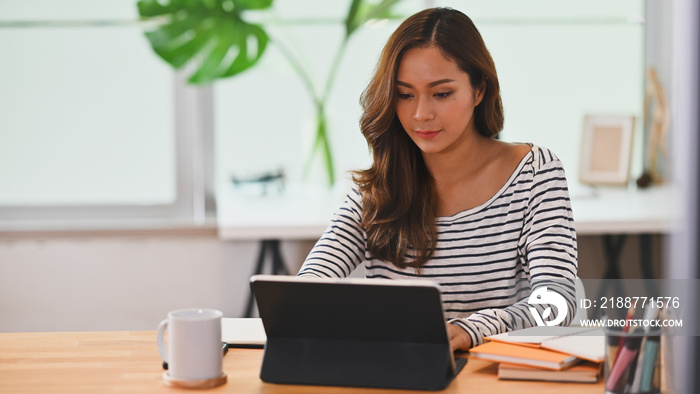 Photo of beautiful creative woman using a stylus pen to drawing on computer tablet with keyboard case that putting on working desk while sitting in orderly living room.