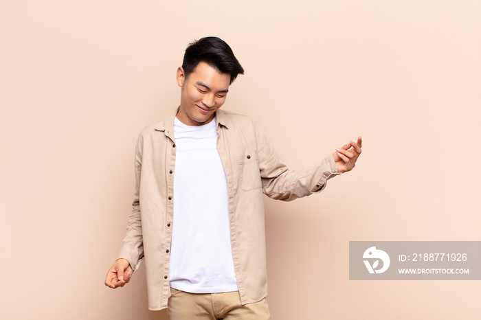 young chinese man smiling, feeling carefree, relaxed and happy, dancing and listening to music, having fun at a party against flat color wall