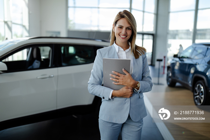 Female salesperson at a car showroom, holding a notes and smiling to the camera.