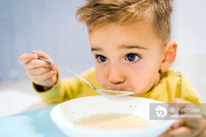 Portrait of small boy child eating soup meal or breakfast having lunch by the table at home with spoon white