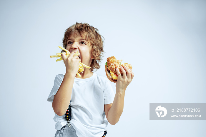 Pretty young curly boy in casual clothes on white studio background. Eating burger with fried potato. Caucasian male preschooler with bright facial emotions. Childhood, expression, fun, fast food.