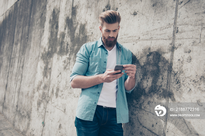 Young serious man in casual clothes leaning on the concrete wall and using smartphone for sending sms