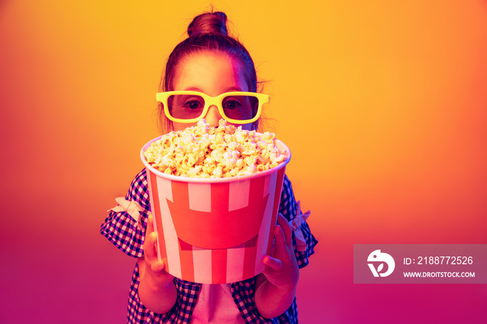 One happy little girl, pupil wearing 3d glasses and holding bucket of popcorn isolated on orange background in neon. Concept of emotions, facial expression, youth, aspiration