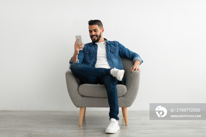 Happy millennial Arab man sitting in armchair with smartphone, texting to friend, sending sms, browsing web