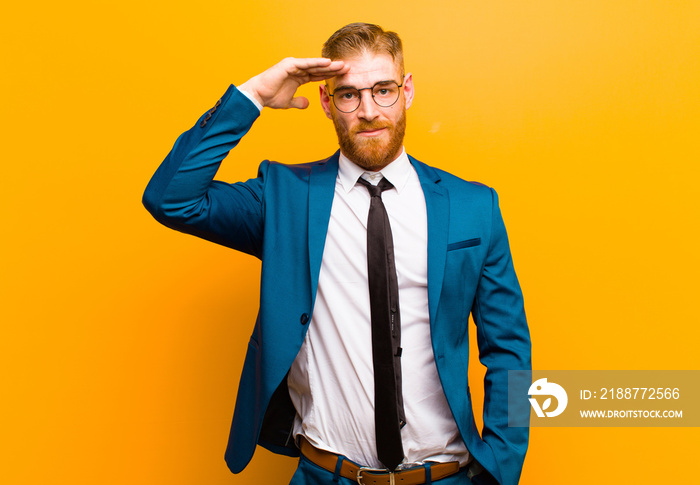 young red head businessman greeting the camera with a military salute in an act of honor and patriotism, showing respect against orange background