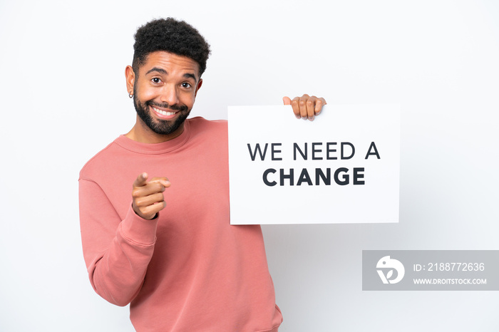 Young Brazilian man isolated on white background holding a placard with text We Need a Change and pointing to the front