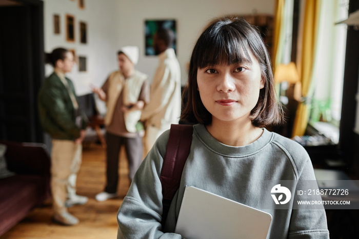 Portrait of Asian student with backpack and textbook looking at camera standing in the classroom with students in the background