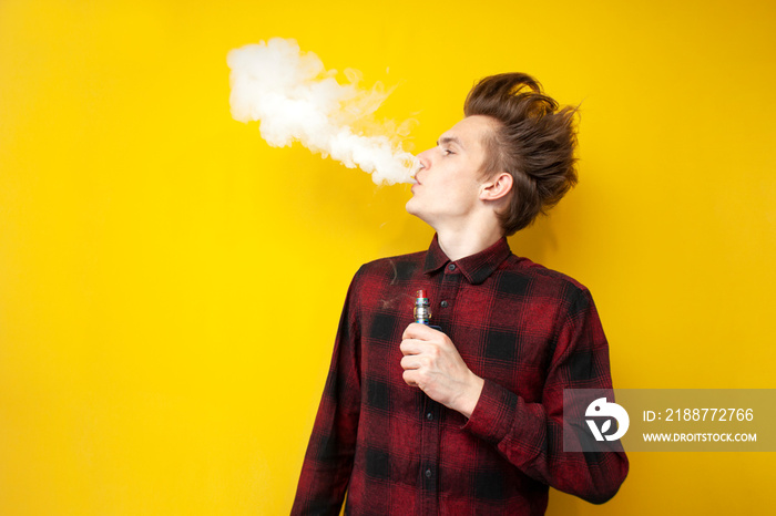 guy hipster smokes an electronic cigarette on a yellow isolated background, a man exhales a cloud of smoke and holds a device for smoking