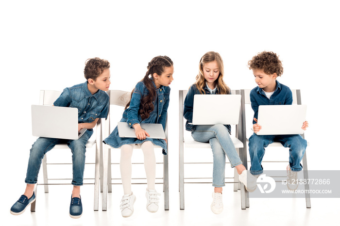 four kids in denim clothes sitting on chairs and using laptops on white