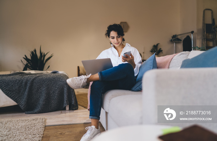 Content woman browsing smartphone and laptop on cozy sofa