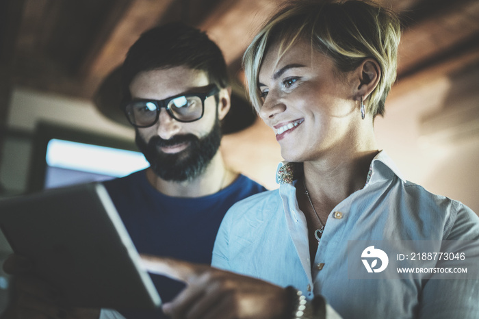 Happy family couple using electronic tablet in livingroom at home.Bearded man in eye glasses making conversation with young blonde woman at night. Blurred background.