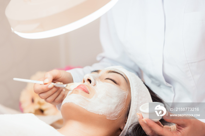 Close-up of the face of a young woman relaxing under the gentle touch of the specialist, applying on her cheeks white facial mask with rejuvenating effects