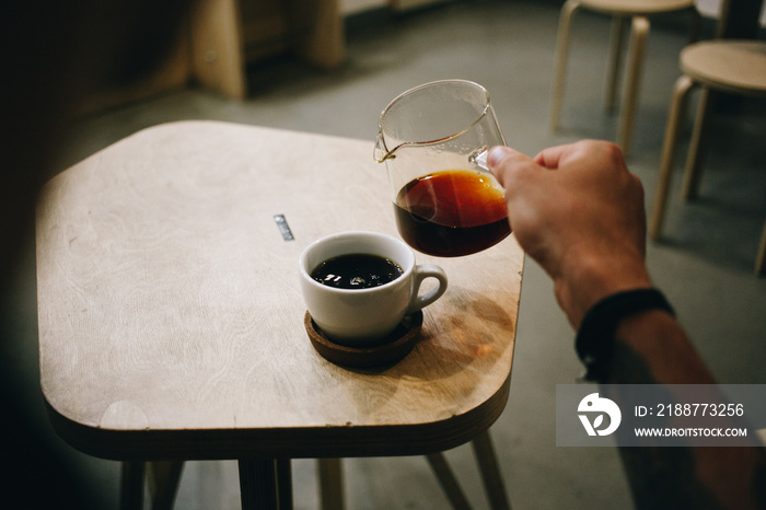 Cinematic vintage photo behind shoulder of man with arm tattoos pour filter coffee from glass jar into cup at specialty coffee shop or cafe. fancy caffeine drink addiction and millennial trend