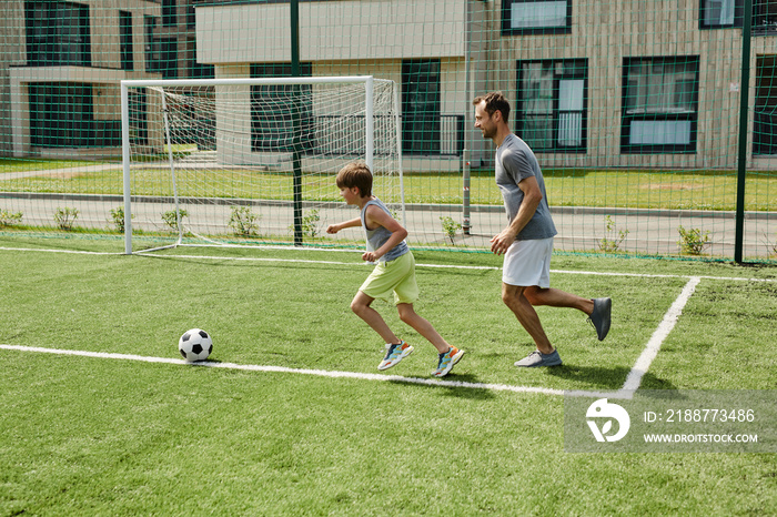 Side view portrait of father and son playing football together in outdoor court and running across grass