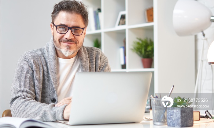 Happy older man working online with laptop computer at home sitting at desk. Home office, browsing internet, study room. Portrait of mature age, middle age, mid adult man in 50s.