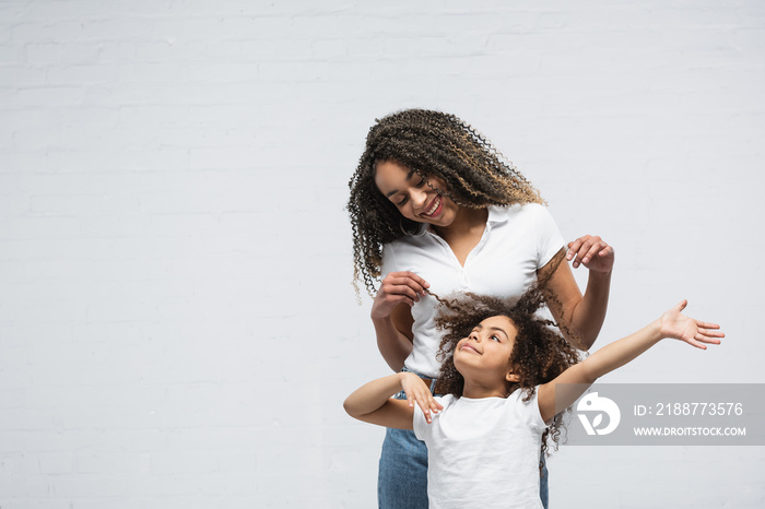 joyful woman touching hair of curly african american daughter on grey