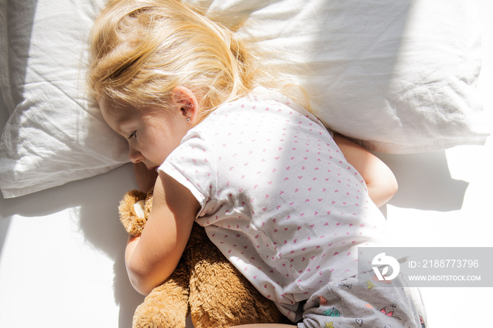 Little girl lies hugging a soft toy bear on a white bed. Top view, flat lay