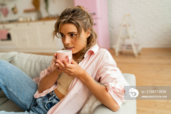 Graceful woman in casual clothes resting un sofa in cozy  living room. Holding cup of  tea.