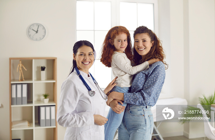 Portrait of cheerful family doctor with woman holding her little daughter together looking at camera. Happy beautiful young mother and pretty child with friendly pediatrician at modern clinic for kids